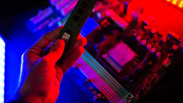 Hand holds RAM module above a computer motherboard illuminated by vibrant red and blue lighting in a dark setting