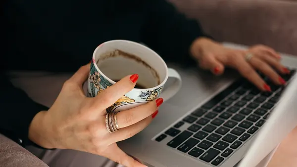 Close-up of a woman with red nail polish holding a cup of coffee while working on her laptop. (50912436636)