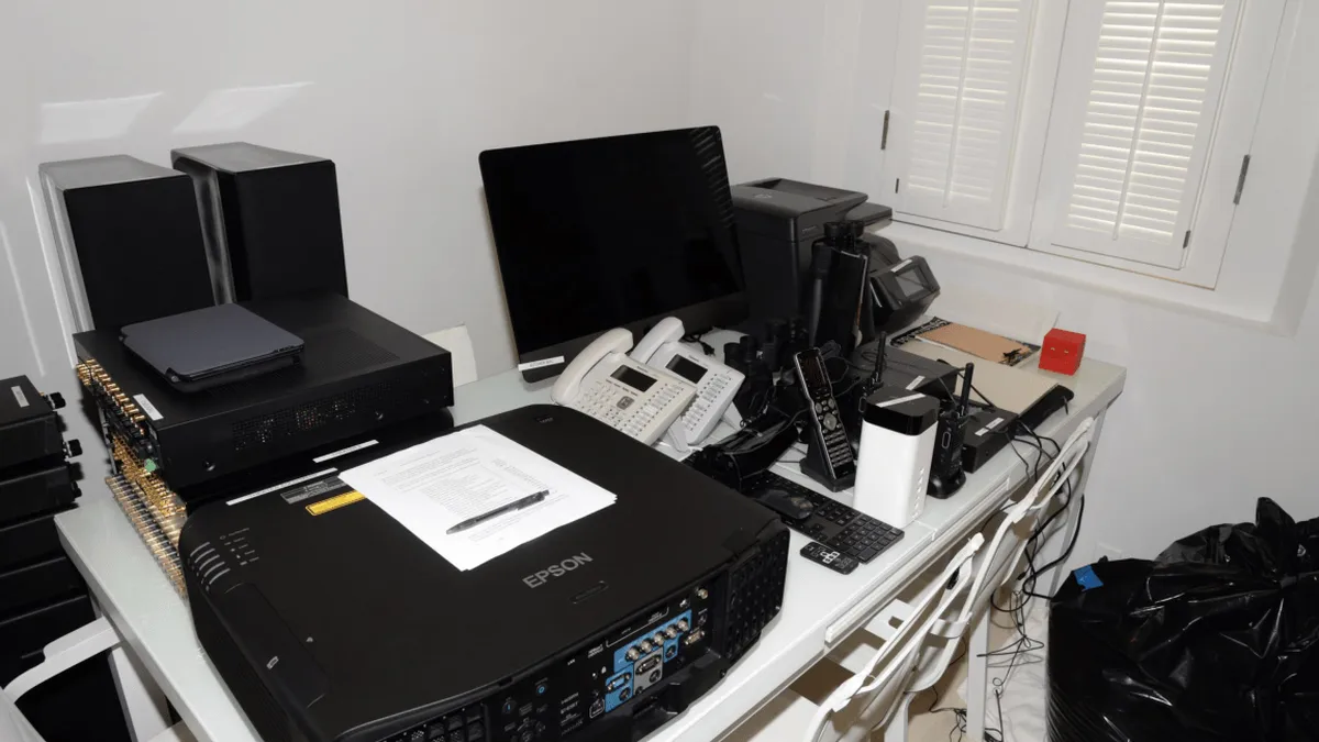 EFTA00001583 - Cluttered office desk with an Epson projector multiple electronic devices a computer monitor and various cables next to a window with shutters