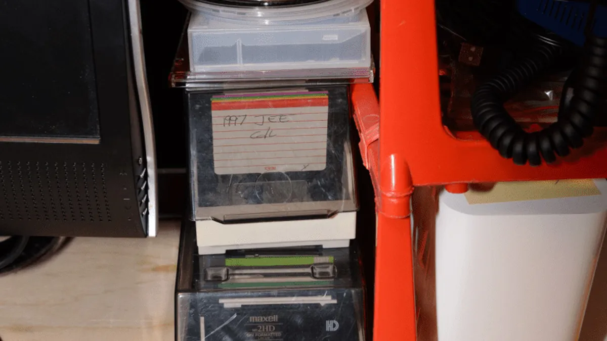 EFTA00001310 - Stack of CDs and storage boxes sits on a wooden floor next to a computer monitor and a red storage rack