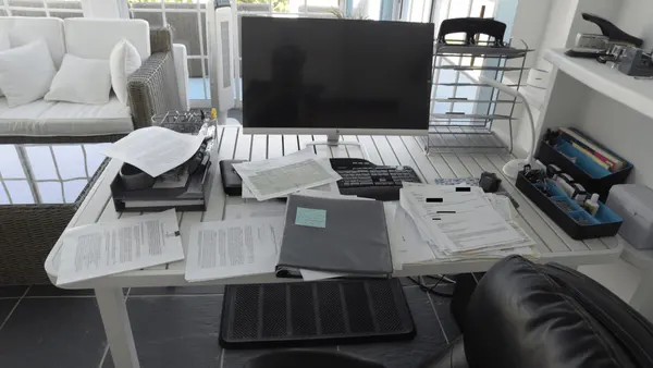 EFTA00003072-Beach-House - Cluttered office desk with papers files and a computer monitor in a bright room with a wicker couch and large windows